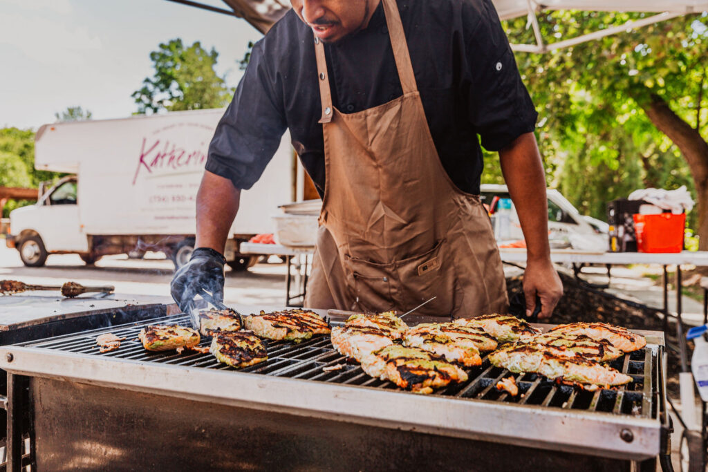 A chef grills marinated chicken outdoors, wearing an apron and gloves, with trees and a truck in the background.