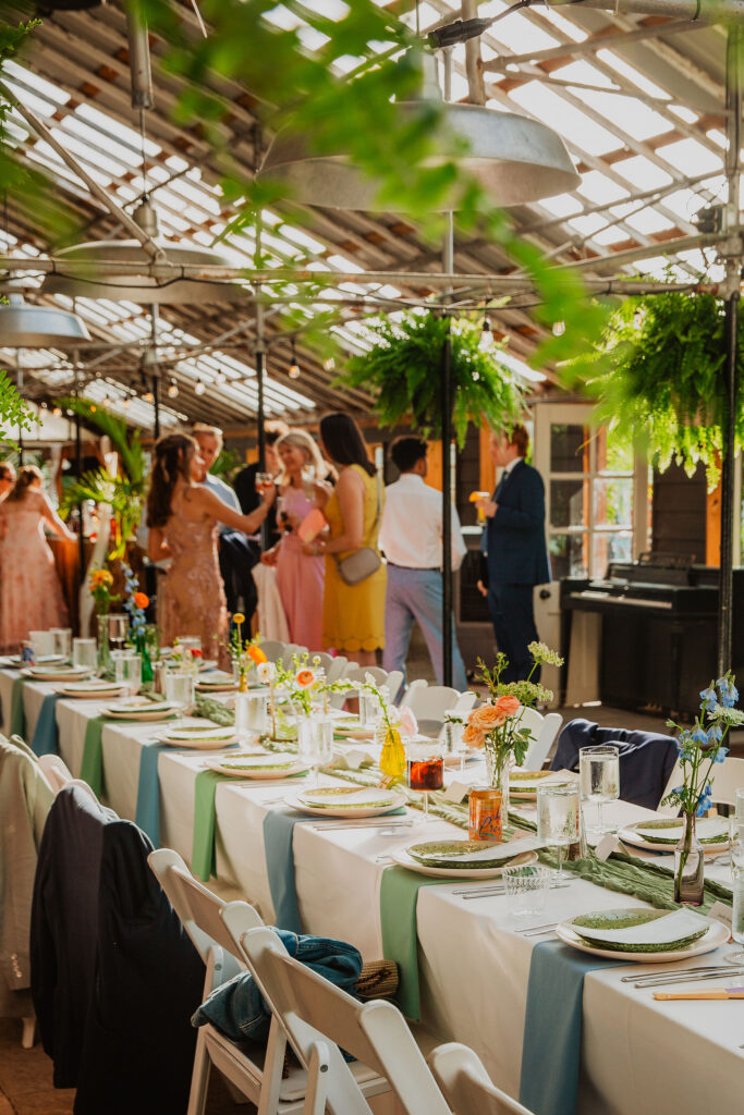 Long banquet table set with plates and glasses; people in formal attire mingling in a sunlit, plant-filled room.