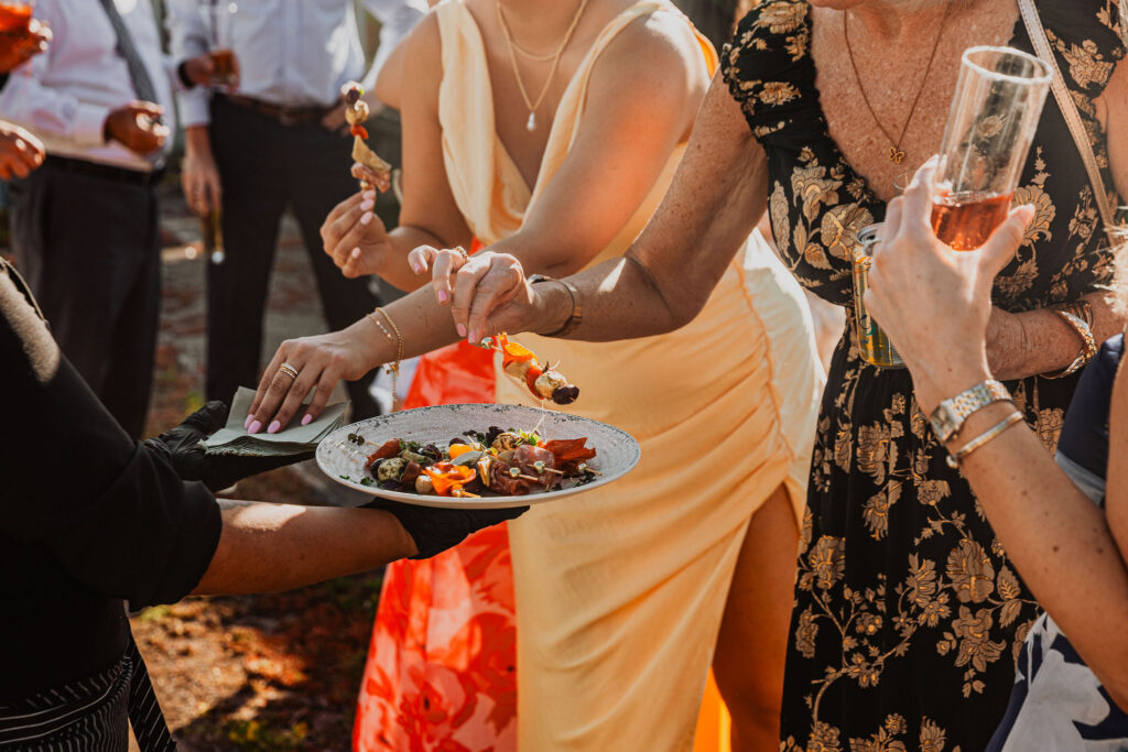 Women in dresses take appetizers from a server holding a plate at an outdoor event; drinks in hand.