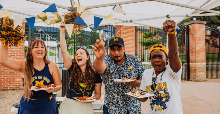 Four people in University of Michigan gear cheer and hold plates of food under a tent at an outdoor event.