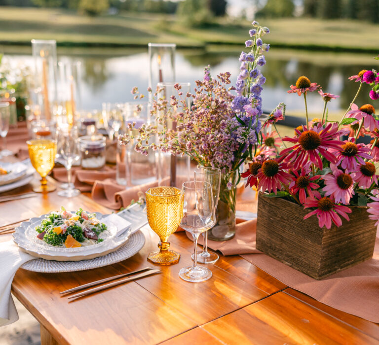 Outdoor table set for a meal with yellow glasses, flower centerpieces, plates of salad, and a lake in the background.
