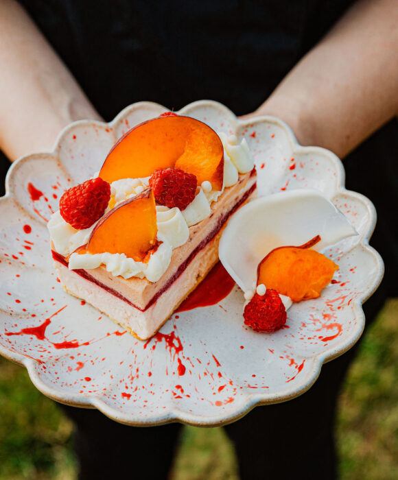 Hands holding a scalloped plate with cake topped with peach slices, raspberries, and whipped cream outdoors.